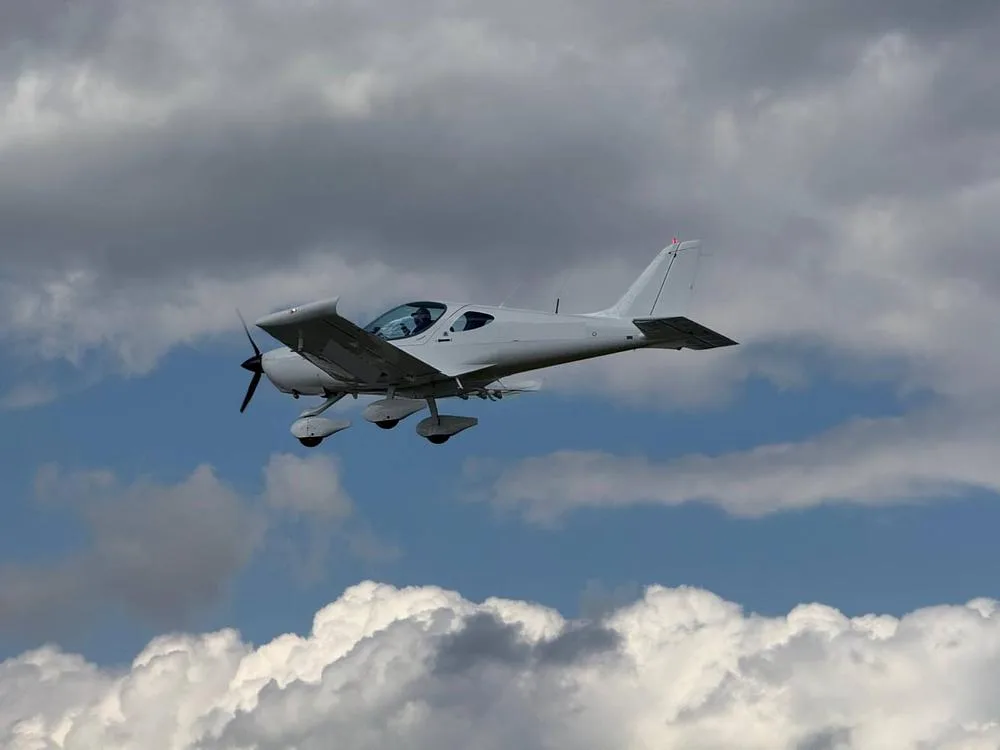 A white Bristell aircraft flying in a cloudy sky, viewed from below.