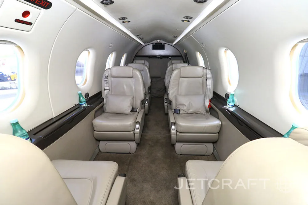 The interior cabin of a Pilatus PC-12 aircraft with light beige leather seats arranged in a club configuration.