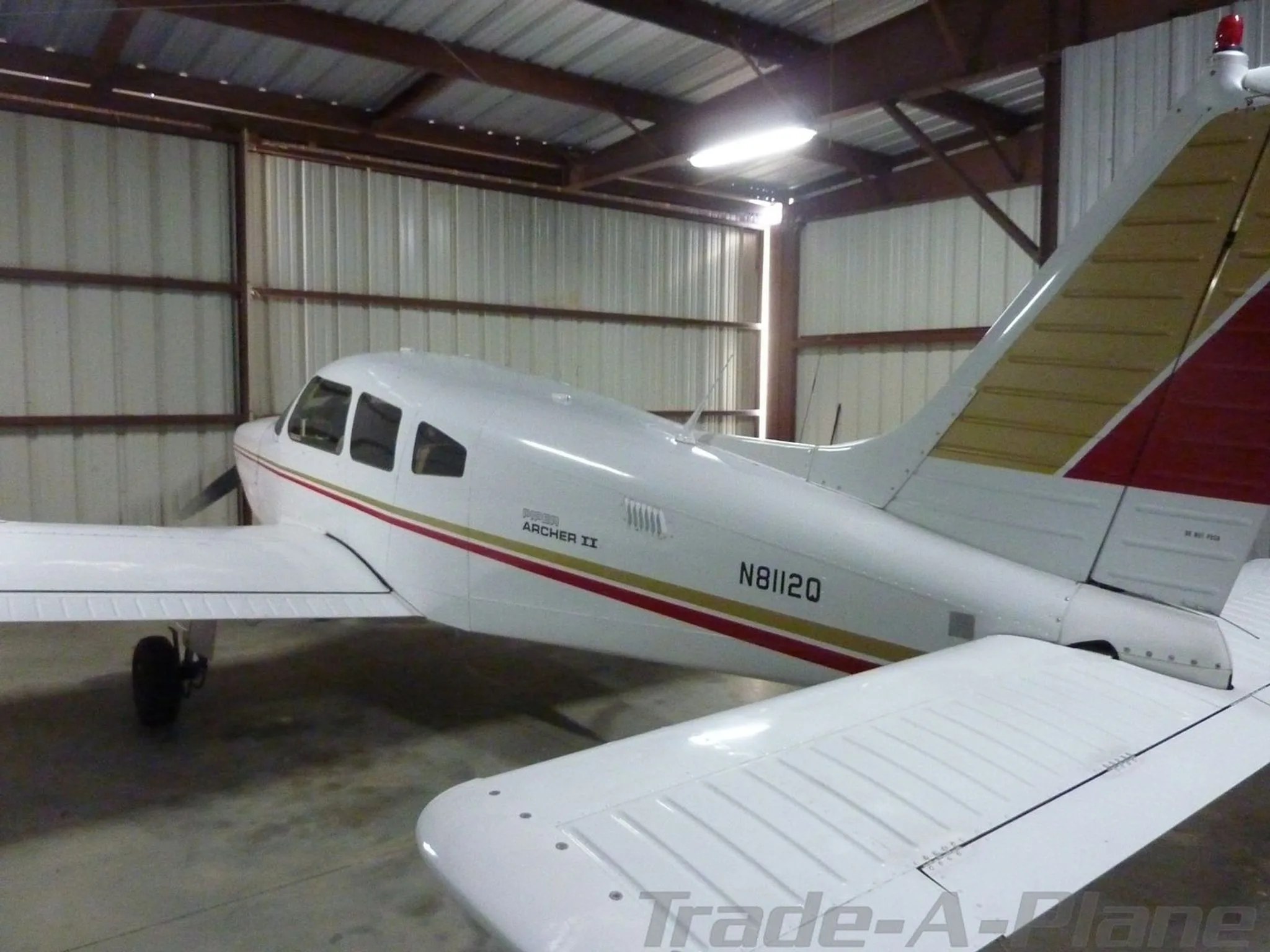 A rear-side view of a white Piper Archer II aircraft with red and gold stripes, registration N8112Q, in a hangar.