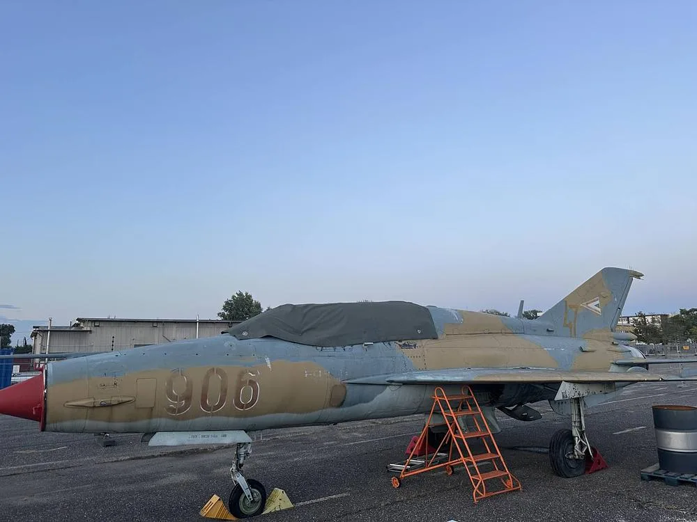 A MiG-21 fighter jet in a two-tone camouflage paint scheme with a red nose cone, parked on an airfield at dusk.
