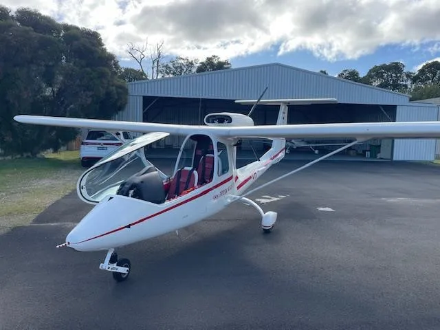 A white light sport aircraft with red stripes and an open canopy is parked on an asphalt apron in front of a hangar.