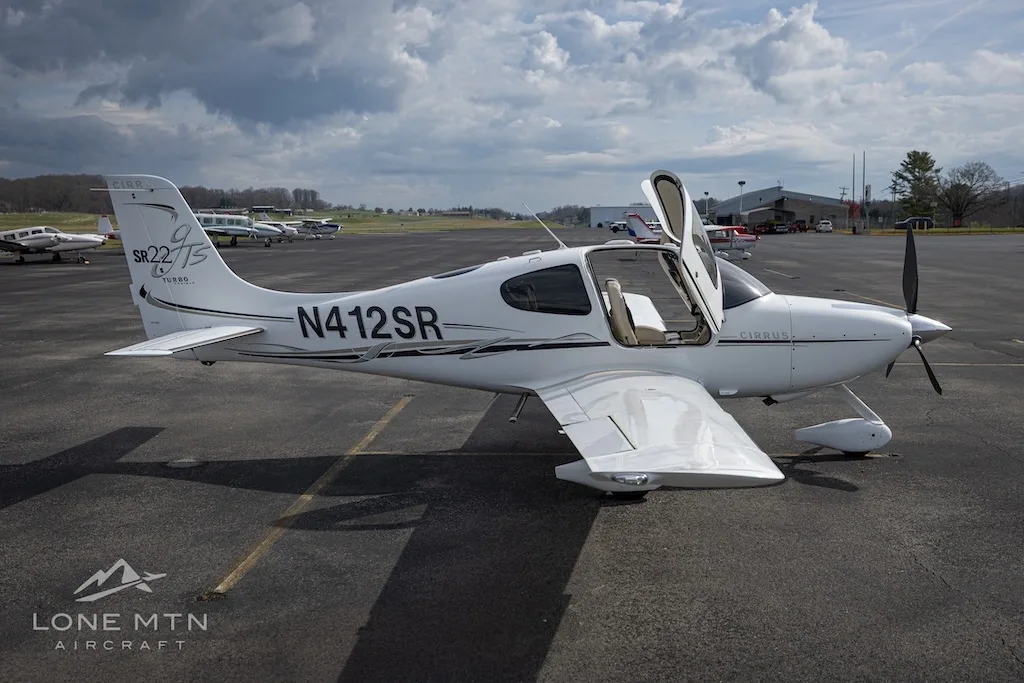 A white Cirrus SR22T aircraft with open doors is parked on an asphalt runway with other planes in the distance.