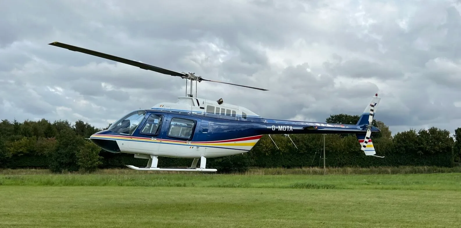 A blue and white Bell 206 helicopter with colorful stripes, registered G-MDTA, hovers over a green field under a cloudy sky.