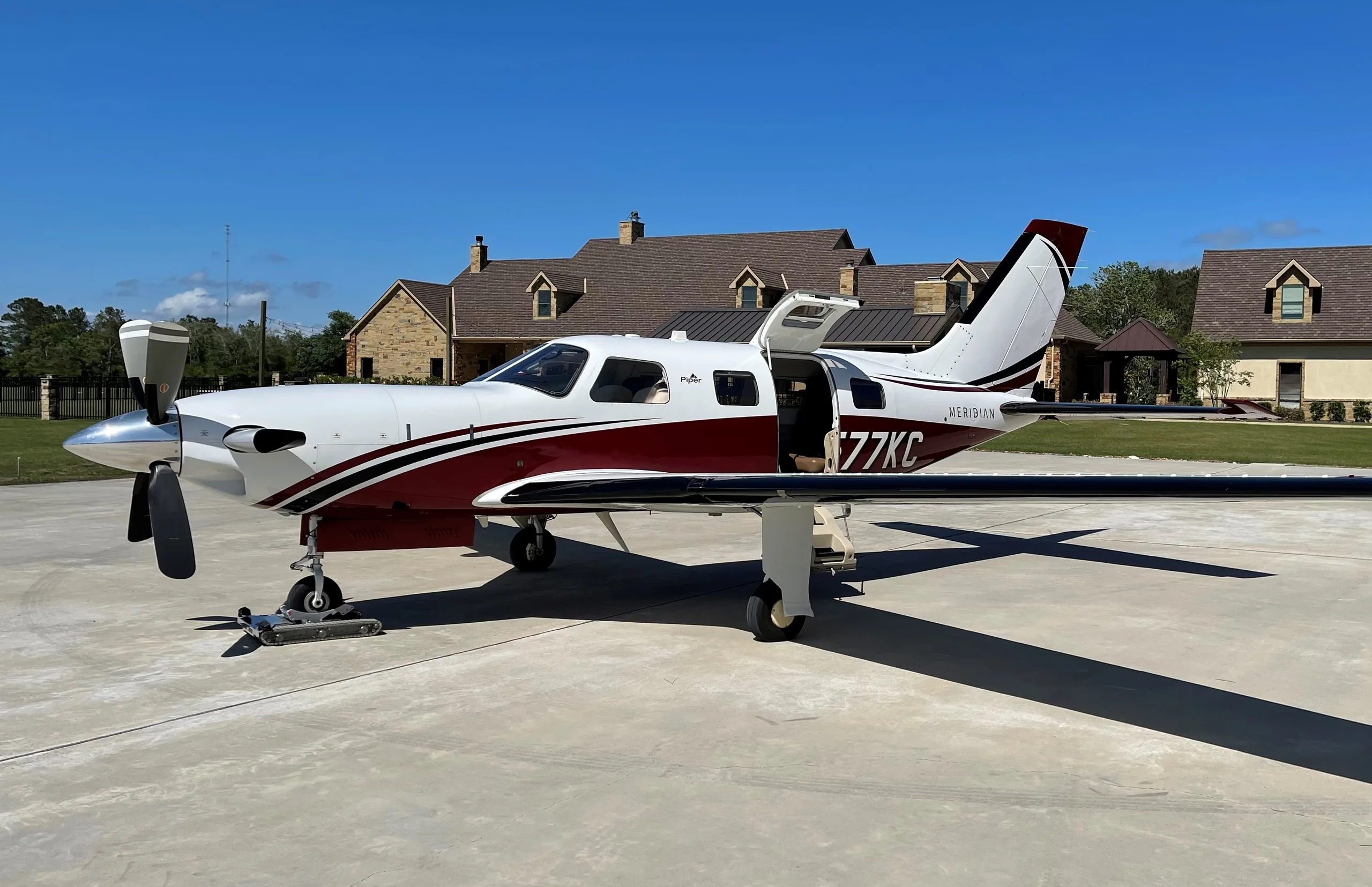 A white Piper Meridian aircraft with red and black stripes, registration N77KC, parked on a concrete pad with its passenger door open, in front of a large house under a clear blue sky.