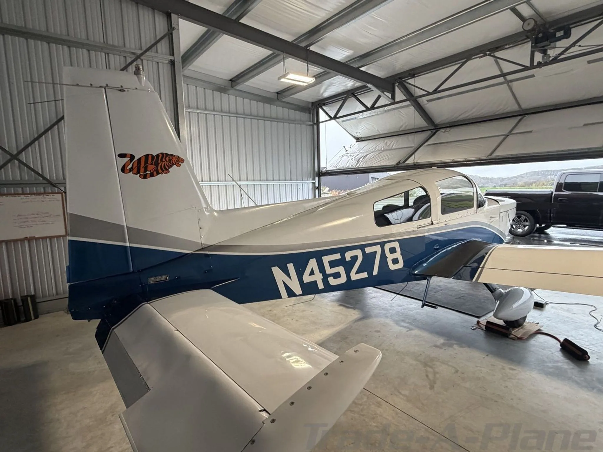Rear-left view of a white and blue Grumman AA-5B Tiger N45278 in a hangar, with a truck visible outside.