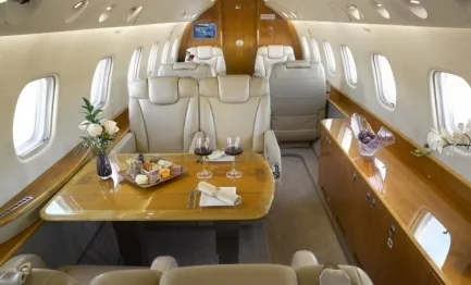 A luxurious aircraft cabin features cream leather seats, a polished wooden table set with food and wine, and large windows.