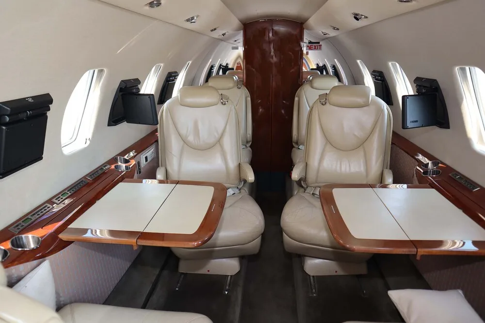 The interior cabin of a business jet featuring beige leather seats, fold-out wooden tables, and a dark wood partition at the rear.