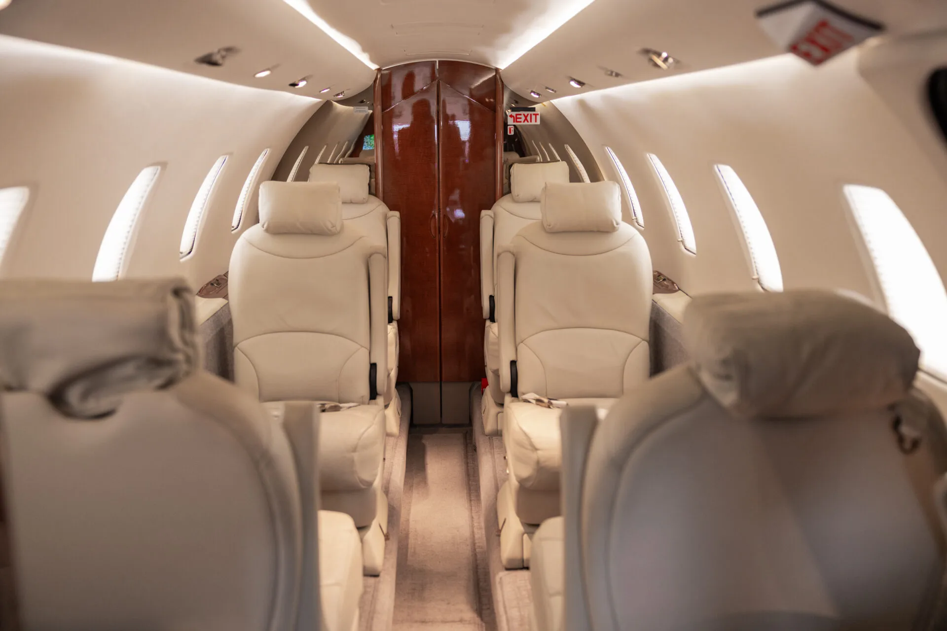 A view down the aisle of a private jet cabin with rows of beige leather seats and an "EXIT" sign visible.