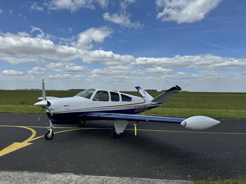 A white and blue Beechcraft Bonanza aircraft (N510CH) parked on an asphalt runway with green fields and a cloudy sky.