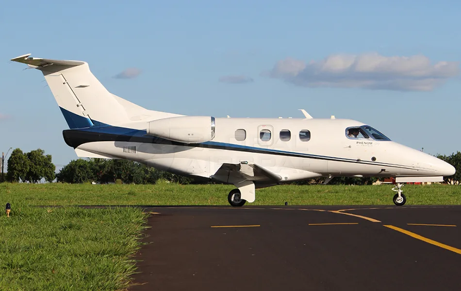 A white Embraer Phenom 300 business jet with a blue and black stripe is seen from the side on an airfield.