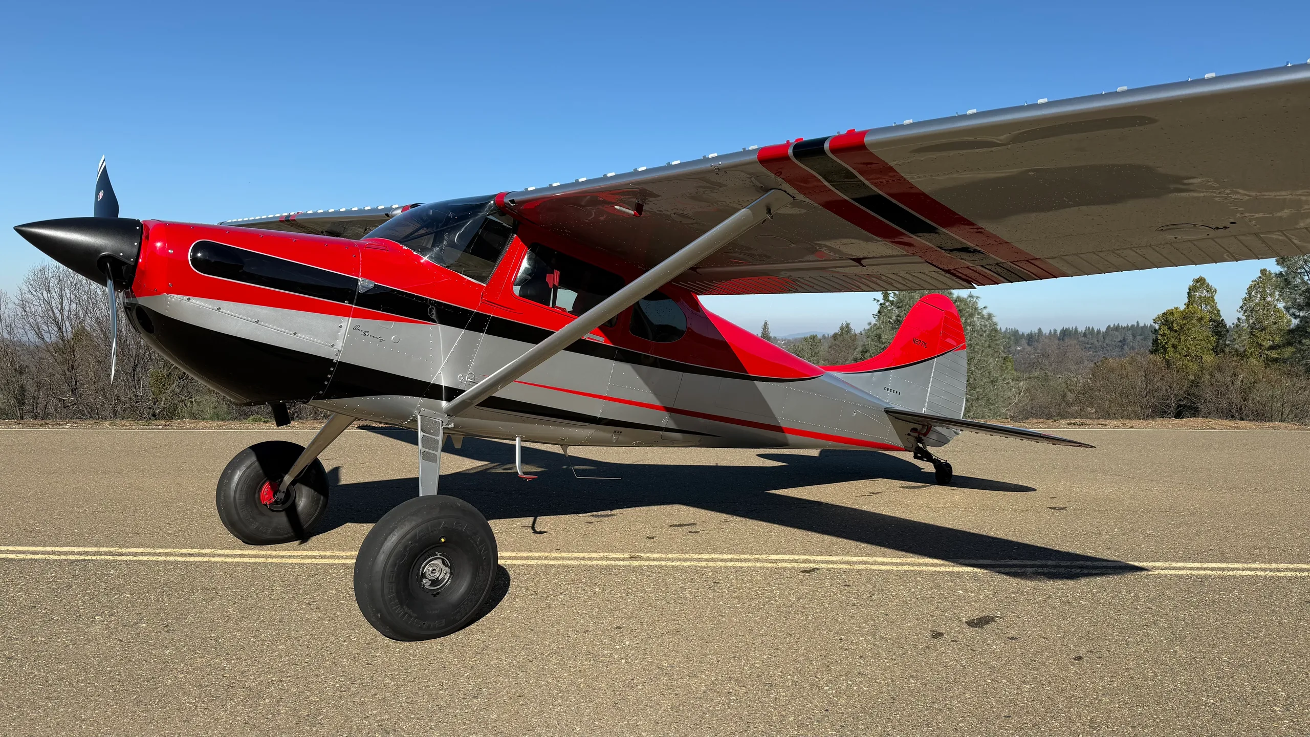 A red, black, and silver Cessna 170 aircraft with large bush tires parked on an asphalt surface under a clear blue sky.