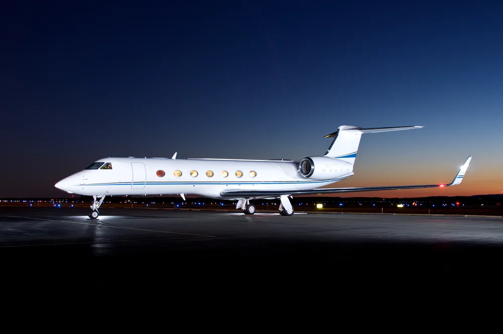 A white Gulfstream G450 business jet with a blue stripe on the tarmac at dusk, viewed from the side.