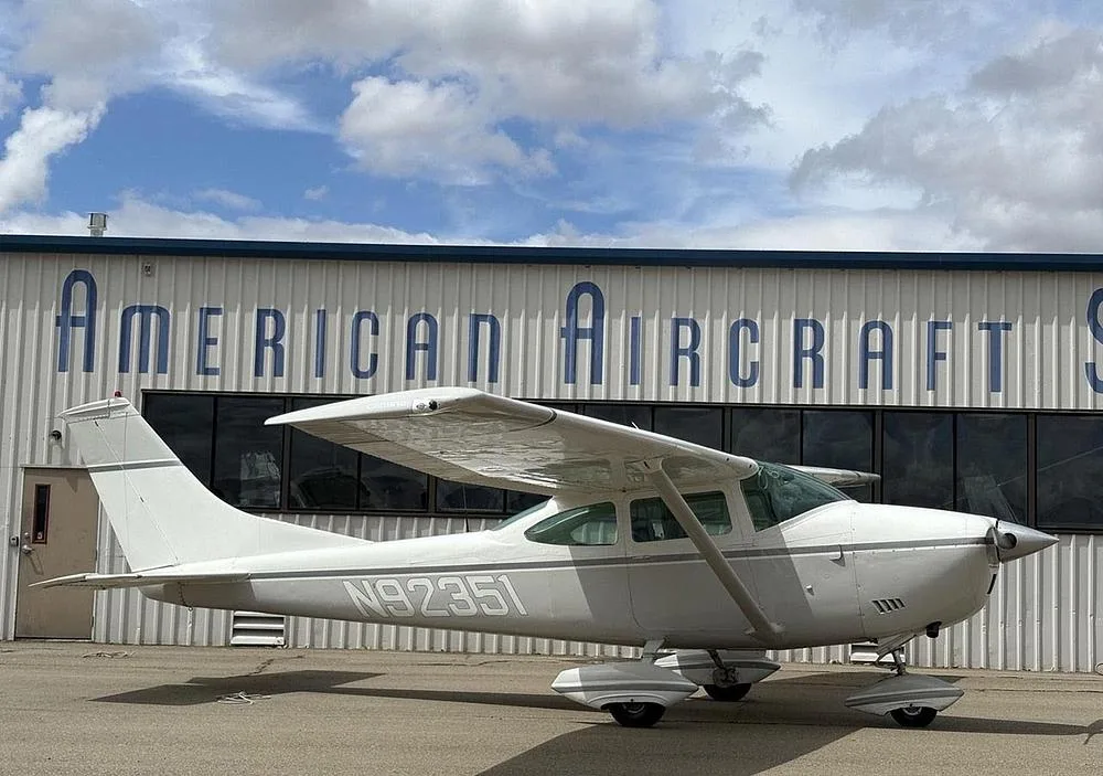 A white Cessna 172 with gray stripes, registration N92351, parked in front of an "American Aircraft" hangar under a cloudy sky.