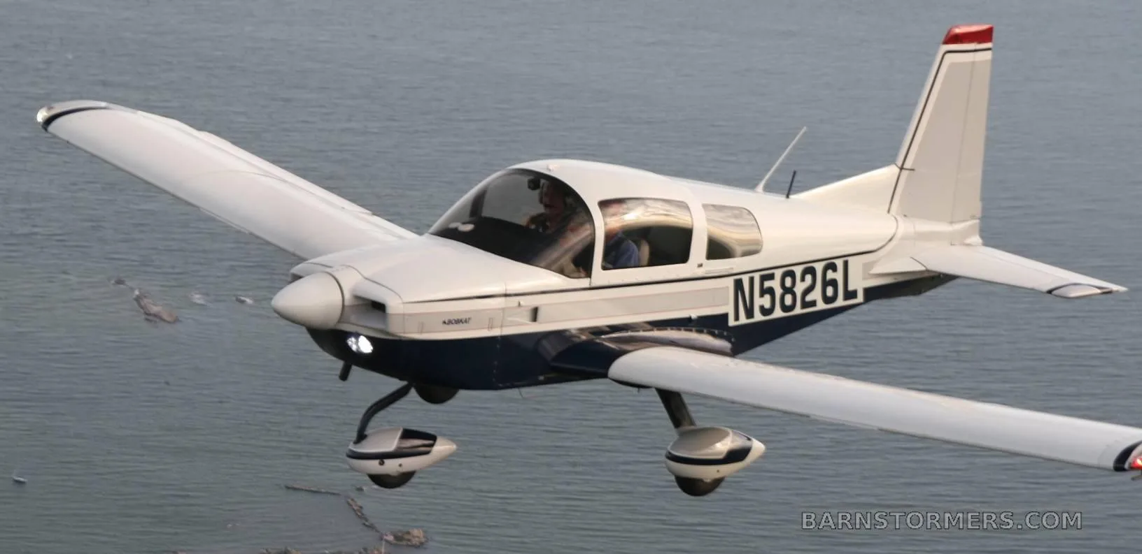 A white and blue Bobcat aircraft (N5826L) with a pilot visible, flying low over water.