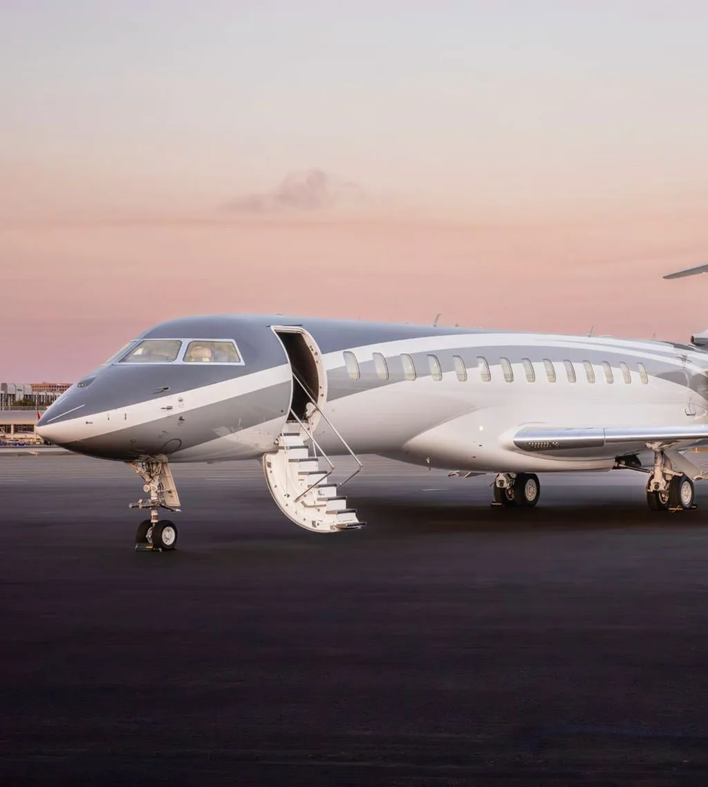 A grey and white private jet, likely a Global 7500, parked on a tarmac at sunset with its main door open and stairs extended.
