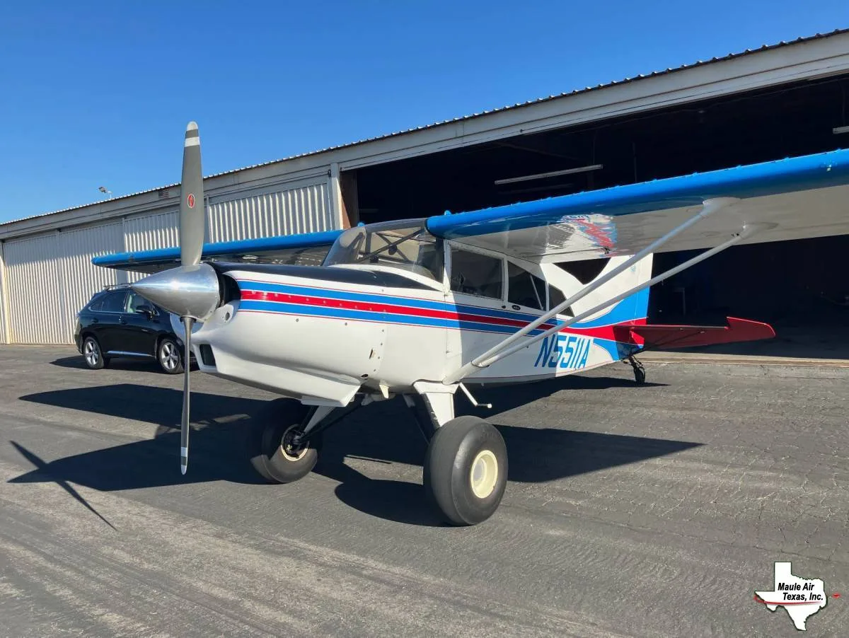 A white Maule aircraft with blue wings and red and blue stripes on the fuselage, registration N5511A, is parked on an airfield in front of a hangar.