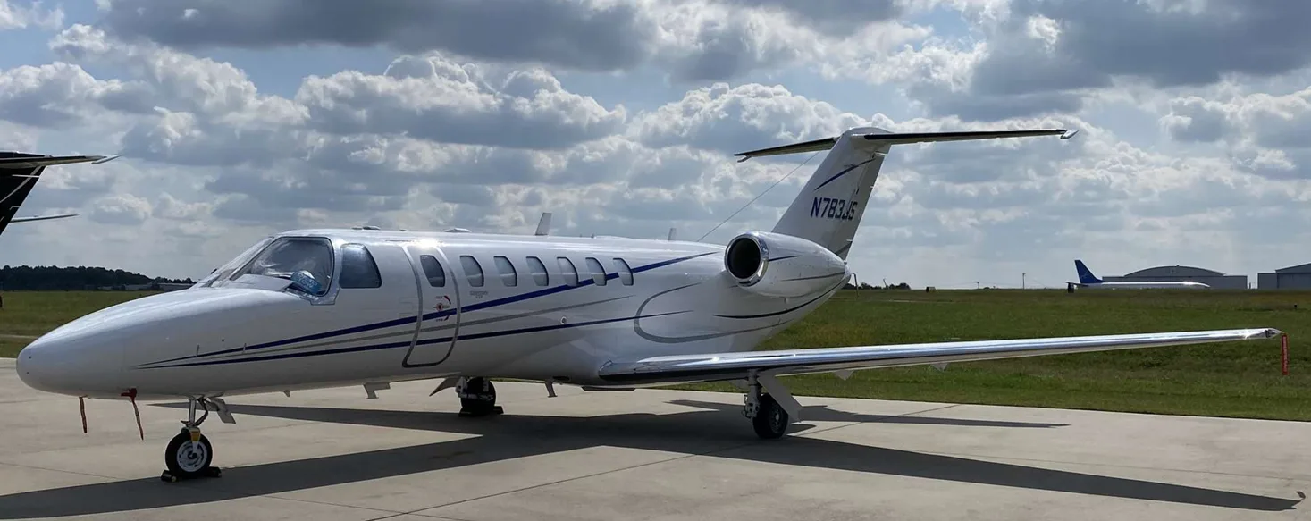 A white Cessna Citation business jet with blue and grey stripes is parked on an airport tarmac under a cloudy sky.