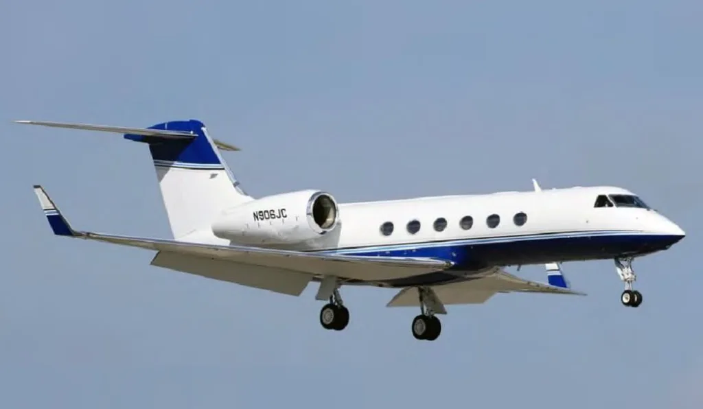 A white Gulfstream G550 with blue stripes, tail number N906JC, in flight with landing gear extended.