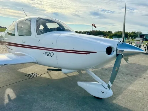 A white Cirrus SR20 aircraft with a red accent stripe is parked on a tarmac, viewed from the front-right.