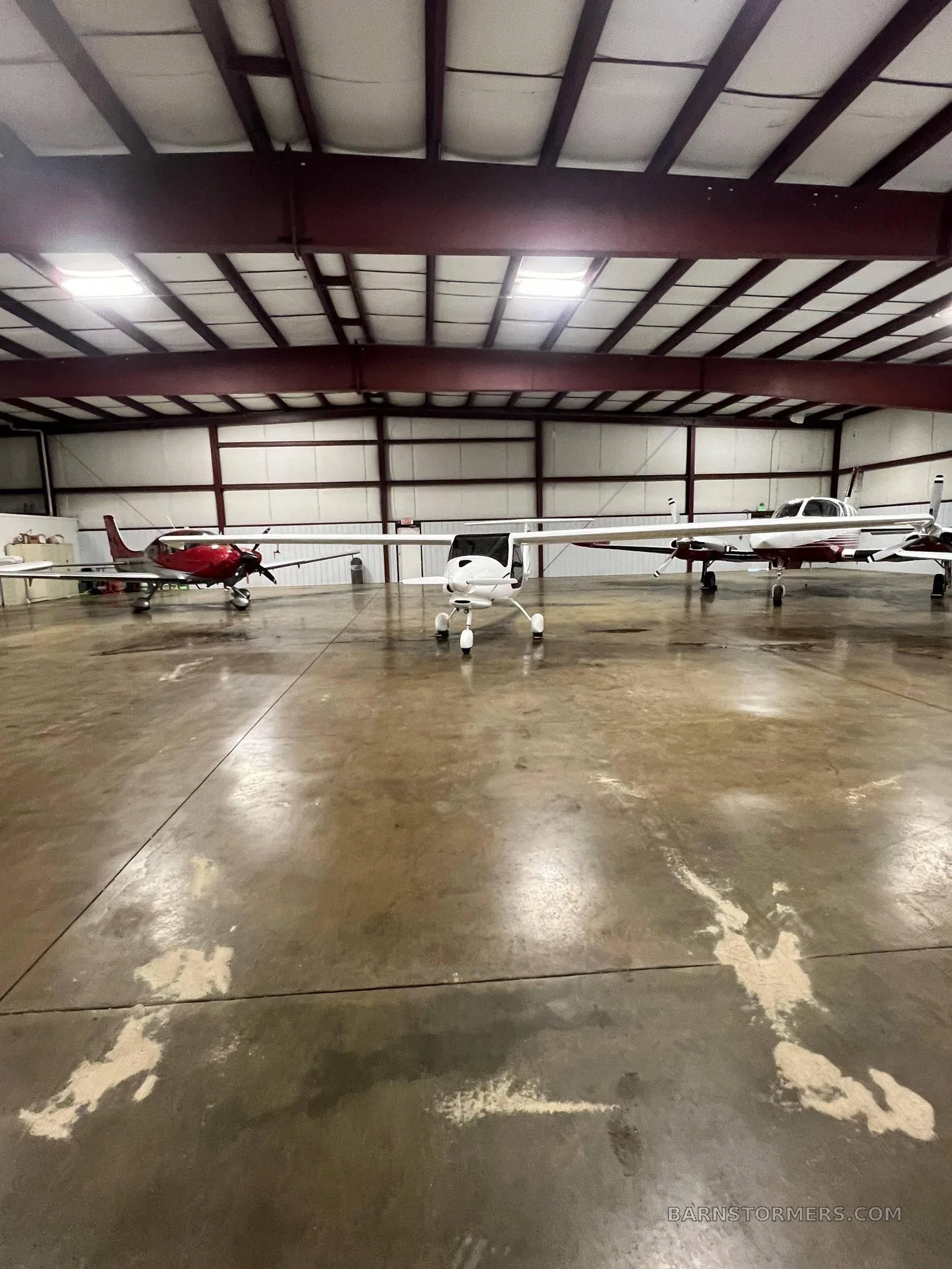 A white Pipistrel Sinus light sport aircraft is centered in a hangar, with two other propeller planes parked on either side.