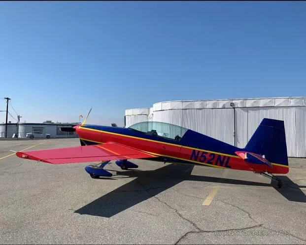 A red and blue aerobatic aircraft with registration N52NL is parked on an asphalt ramp next to a white hangar.