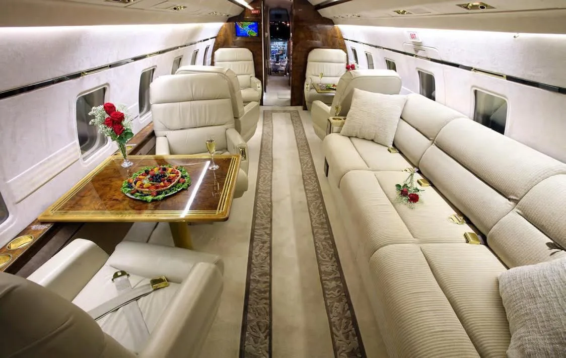 A spacious aircraft cabin featuring cream leather seating, a long sofa, and a dining table with fruit and flowers.