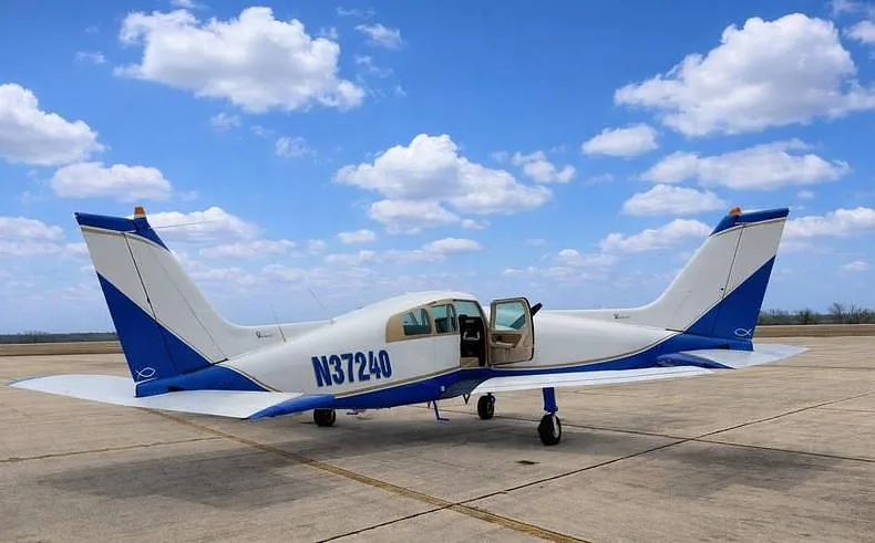 A white and blue Beechcraft Sundowner N37240 with its door open, parked on a tarmac under a partly cloudy sky.