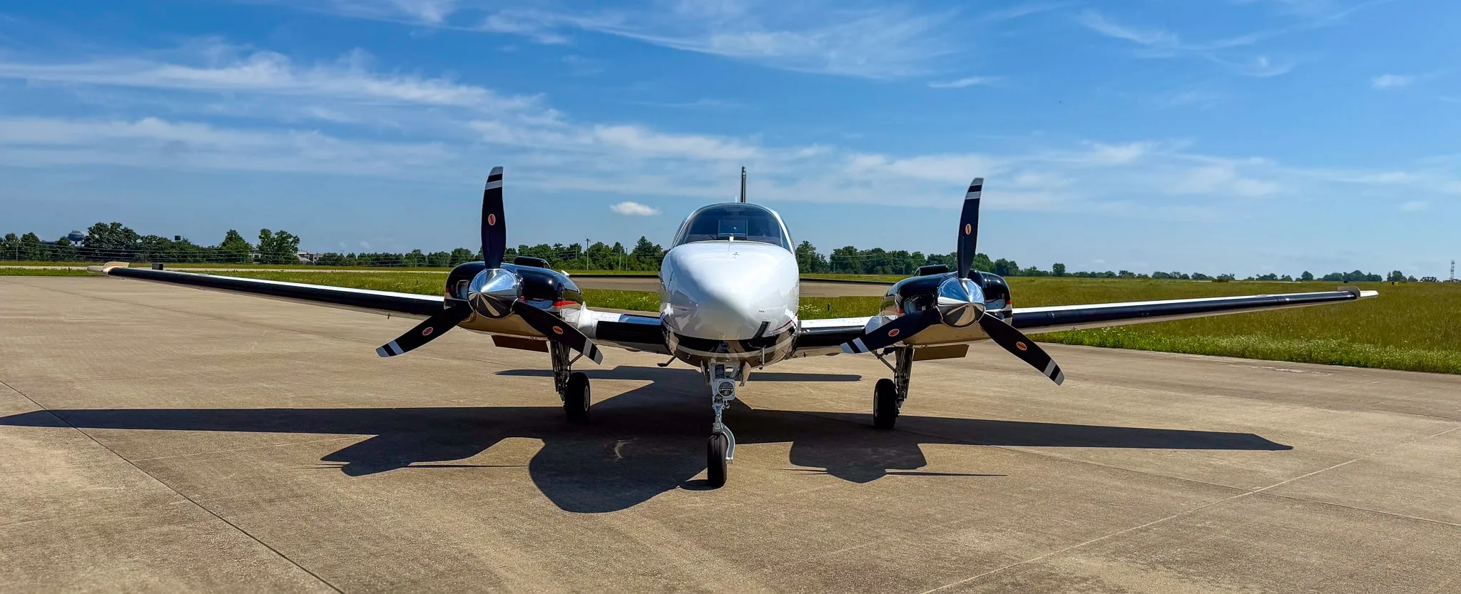 Front view of a white and gold twin-engine Beechcraft Baron G58 on an airfield under a blue sky.