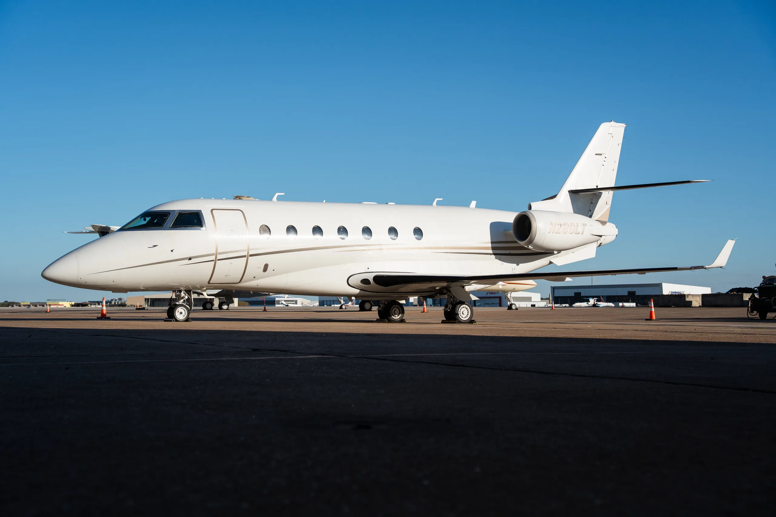A white Gulfstream G280 business jet with gold and black stripes, registration N280LT, parked on an airport tarmac under a clear blue sky.