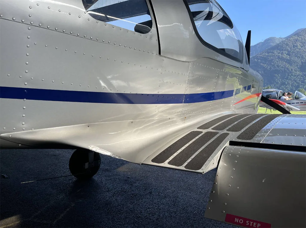 Close-up of the silver fuselage of a Sonaca 200 aircraft showing the blue stripe, step pads, and a "NO STEP" warning.
