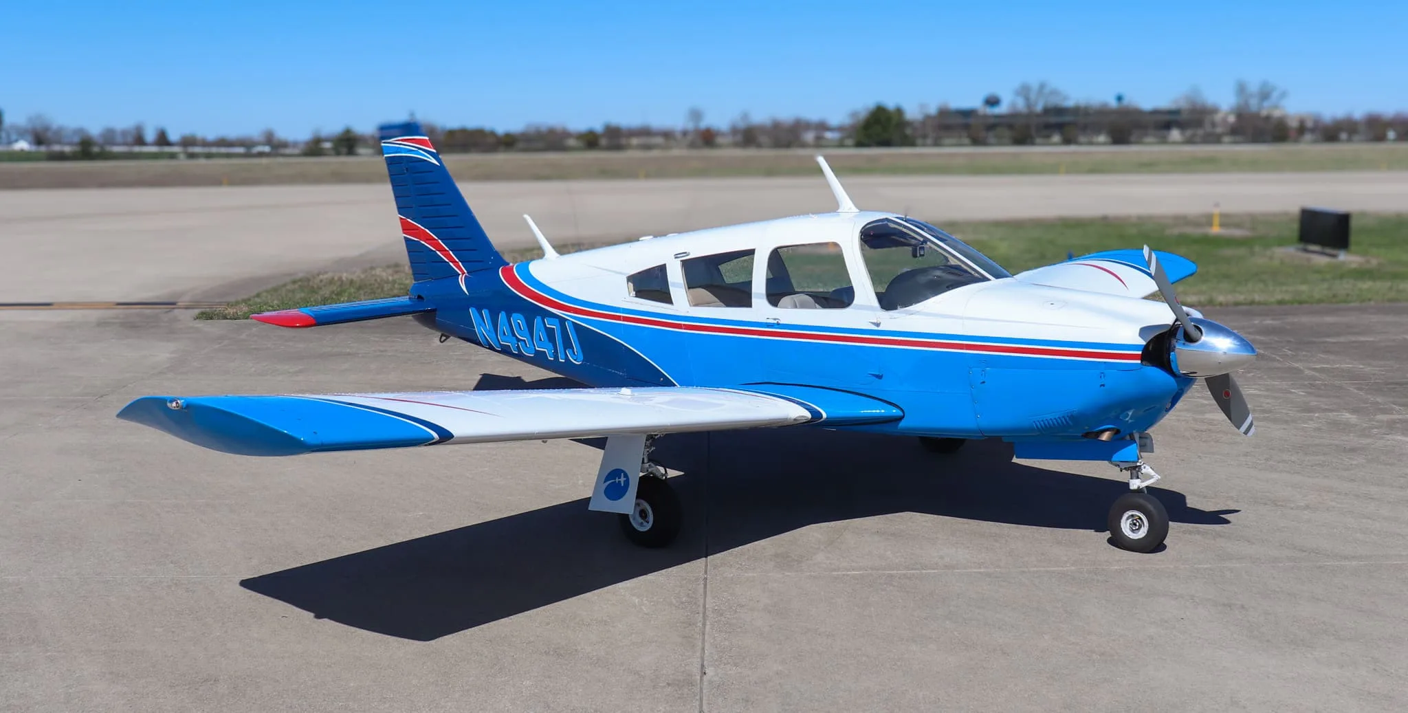 A blue and white Piper Cherokee aircraft with red stripes parked on an asphalt runway under a clear blue sky.