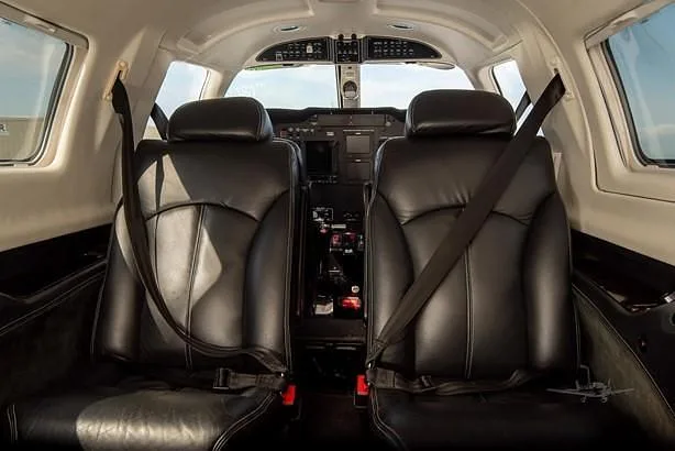 The aircraft cabin shows two black leather passenger seats in the foreground, looking forward towards the cockpit.