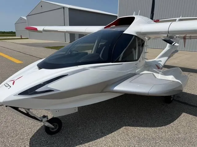A white and silver ICON A5 amphibious aircraft is parked on an airfield with a hangar in the background.