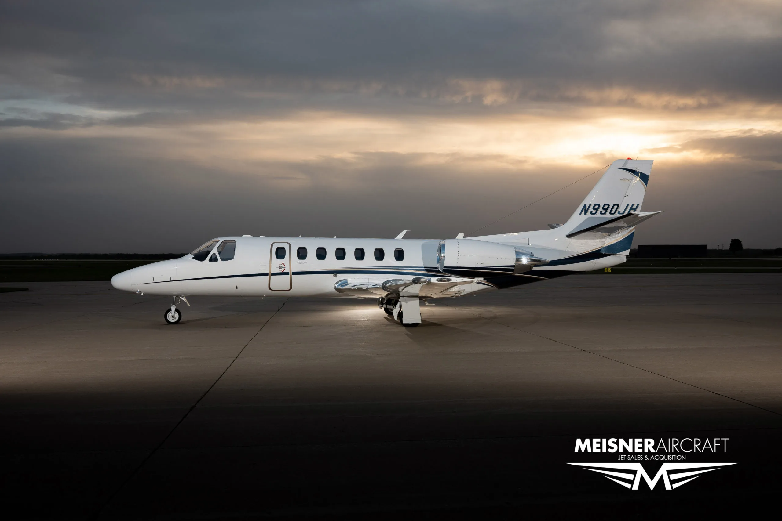 A white Cessna Citation jet with blue and black stripes, registration N990JH, parked on an airfield at sunset, with a 'Meisner Aircraft' logo.