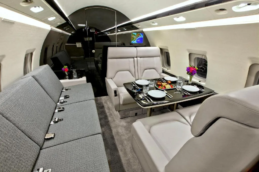 A long view of the private jet's spacious cabin, showing a gray sofa, a dining area with light gray seats, and a galley further back.