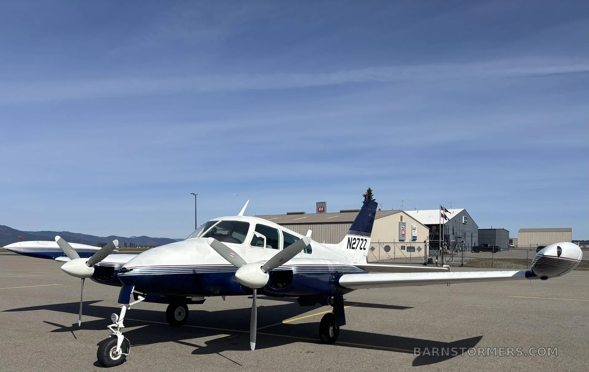 A white and blue Cessna 310I, registration N27ZZ, parked on an airfield with hangars and mountains in the background.
