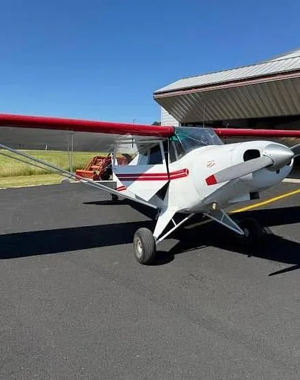 A white aircraft with red wings and stripes parked on an asphalt tarmac outside a hangar.