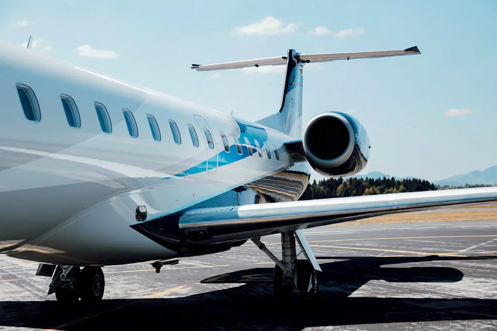 A close-up view of the rear fuselage, engine, and wing of a white and blue Embraer ERJ 145 on an airport tarmac.