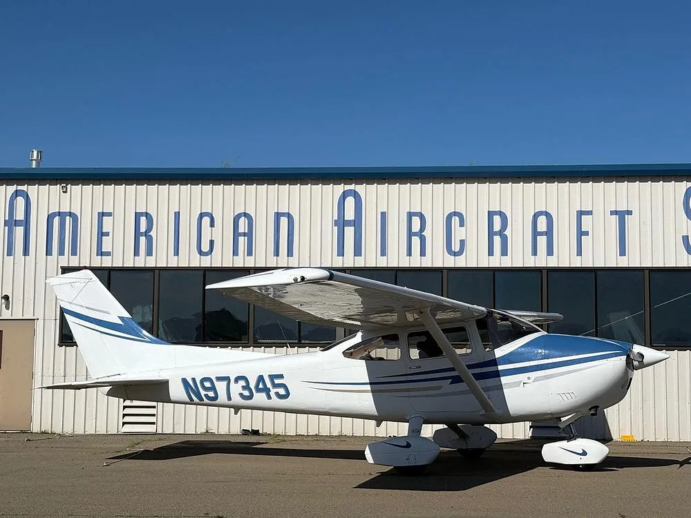 A white and blue Cessna aircraft with registration N97345 parked in front of the American Aircraft Sales building under a clear blue sky.