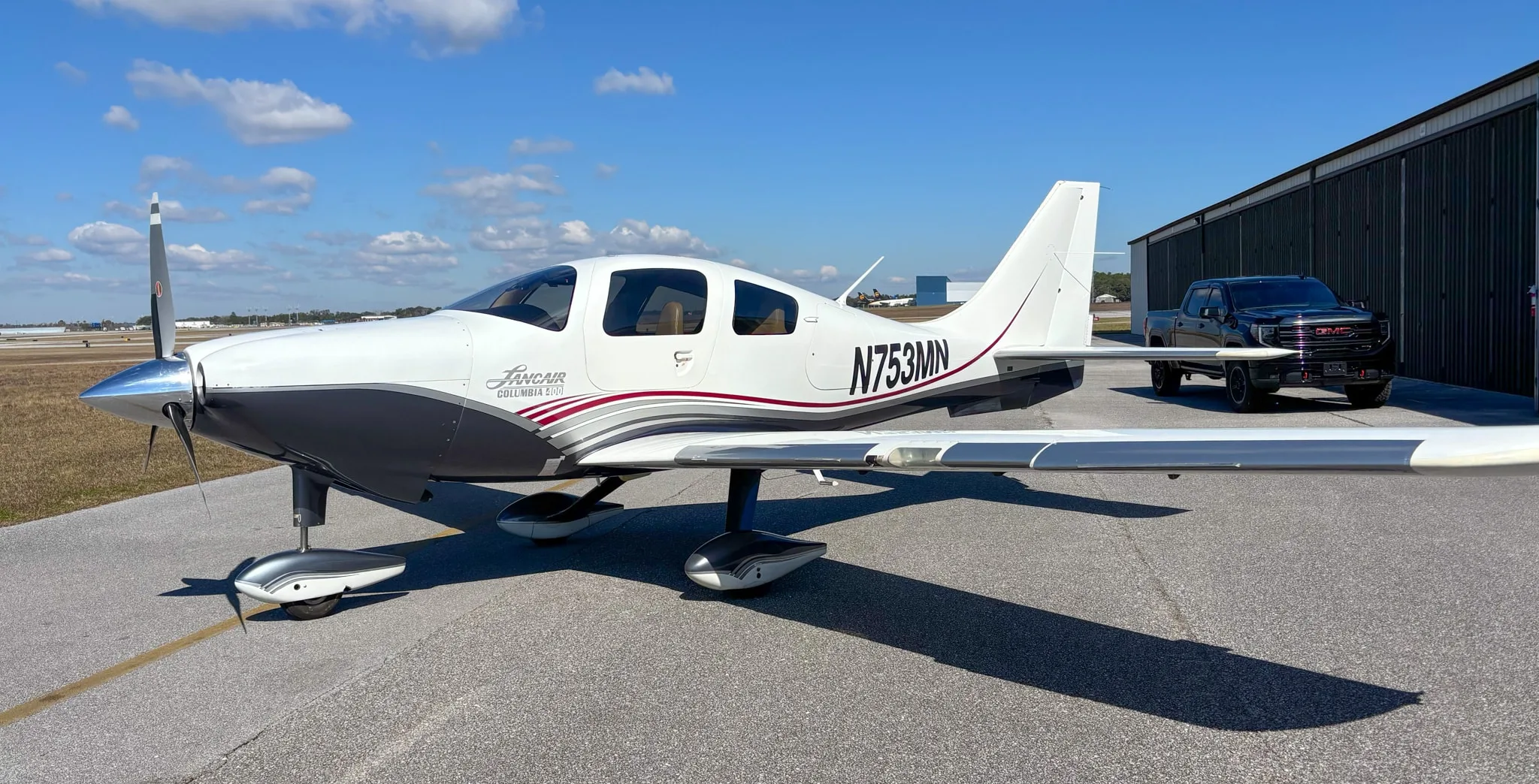 Side view of the white and grey Lancair Columbia 400 N753MN parked on a tarmac next to a hangar.