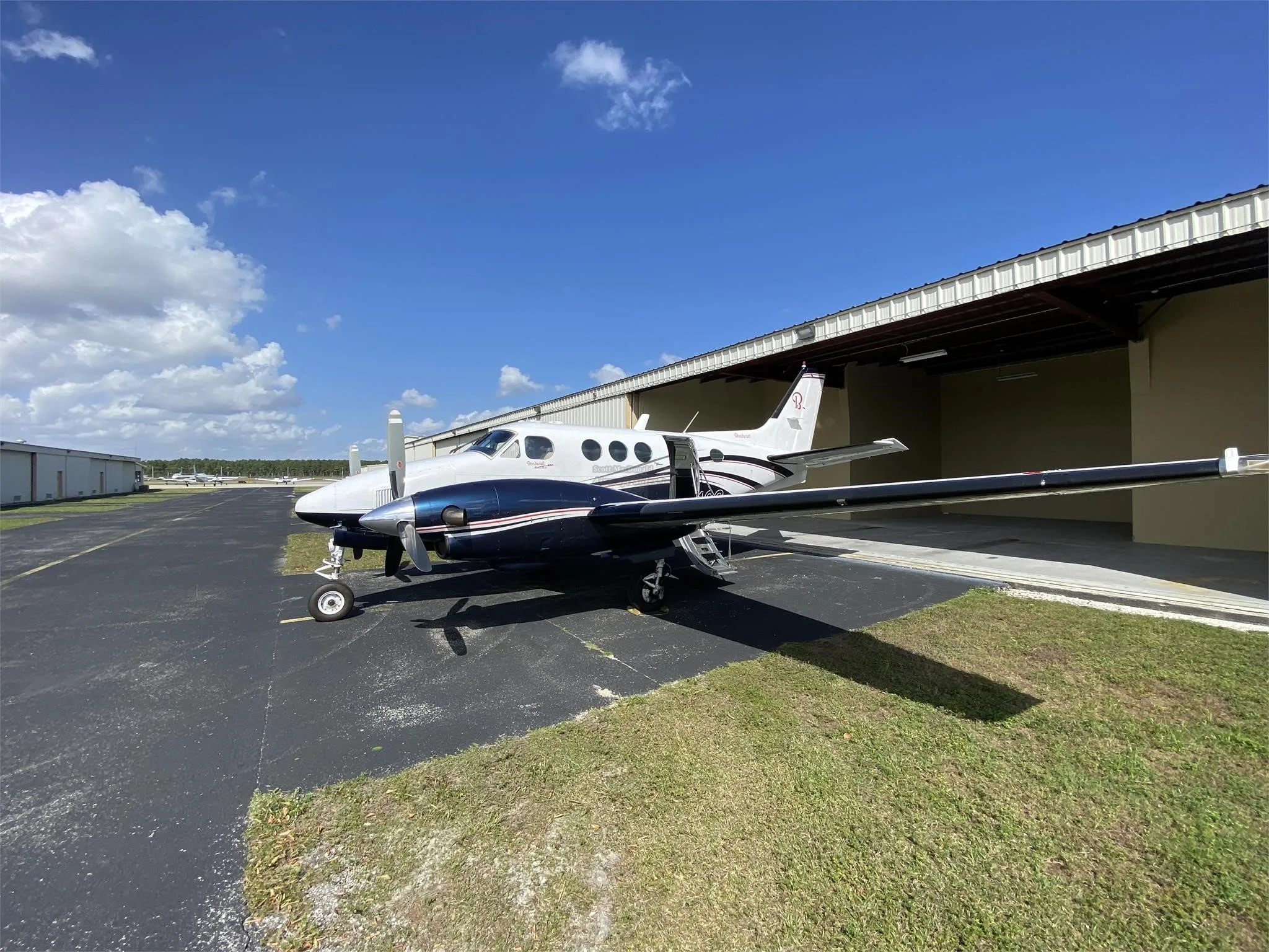 A white and dark blue Beechcraft King Air aircraft parked on an asphalt apron next to a hangar under a partly cloudy sky.