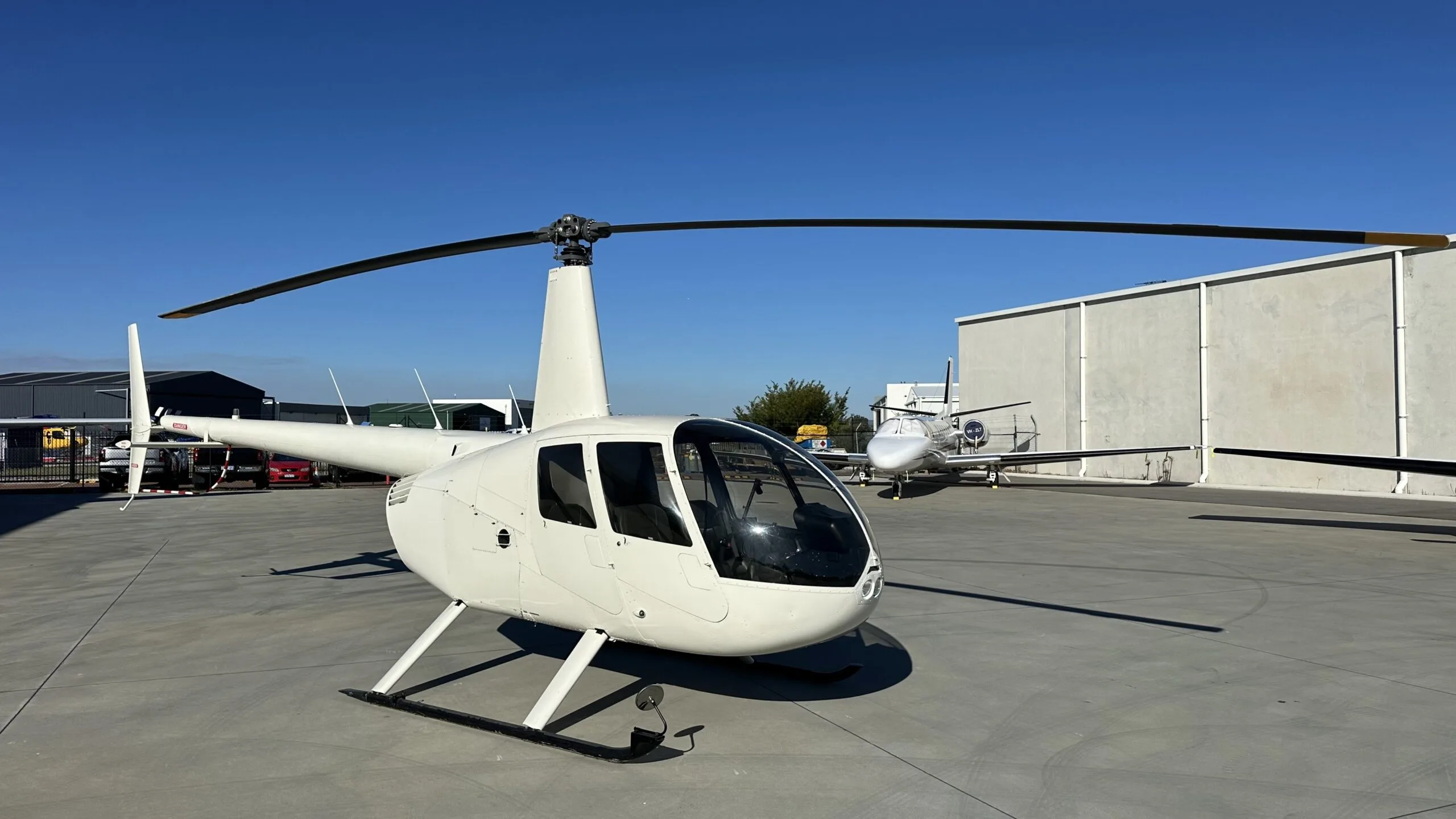 A white Robinson R44 helicopter parked on an airfield tarmac, viewed from the front-right.