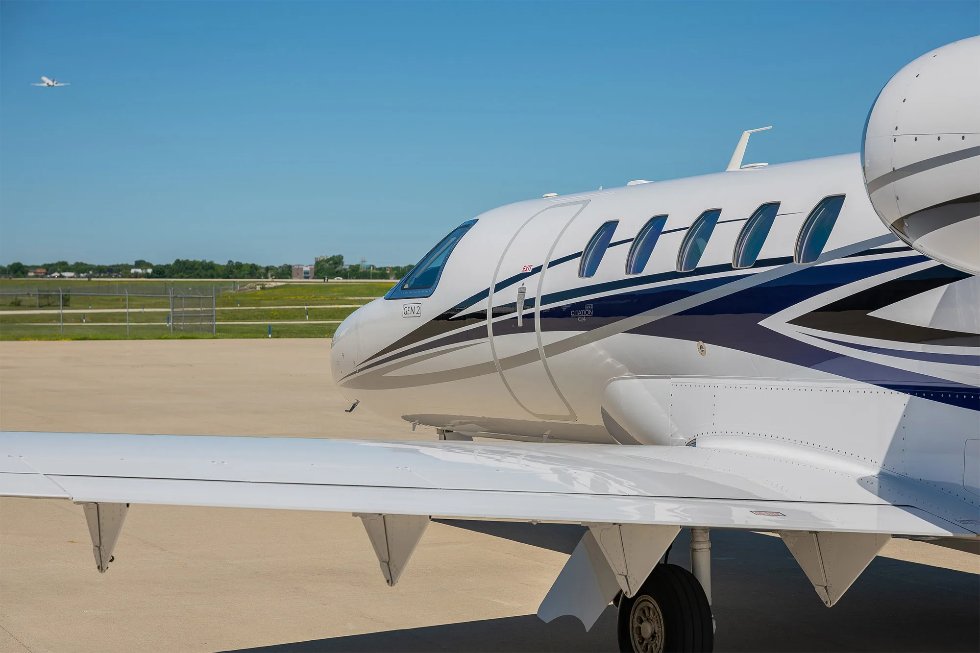 A close-up view of the front fuselage and wing of a white business jet with blue and grey stripes, showing 'GEN 2' text.