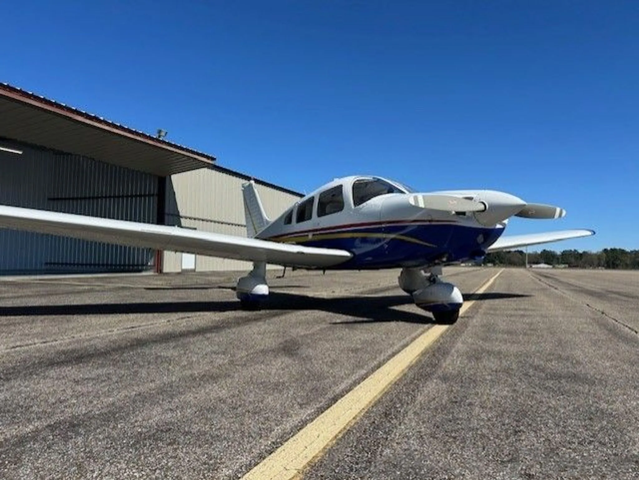 A white and blue single-engine aircraft with red and yellow stripes parked on an airfield in front of a hangar.