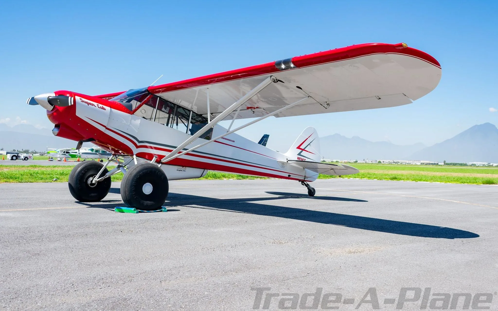 A three-quarter front view of a red and white Super Cub aircraft with N874CC on the tail, parked on an airfield.