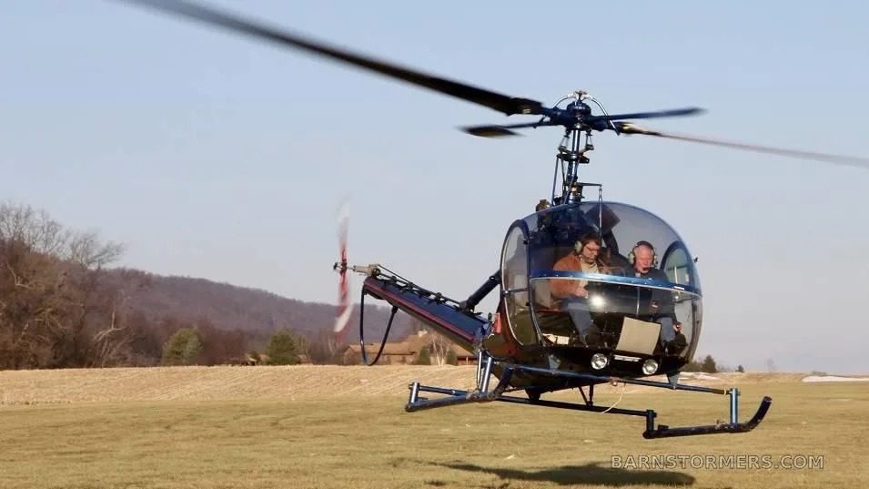 A blue and black helicopter with two occupants flies low over a grassy field on a clear day.