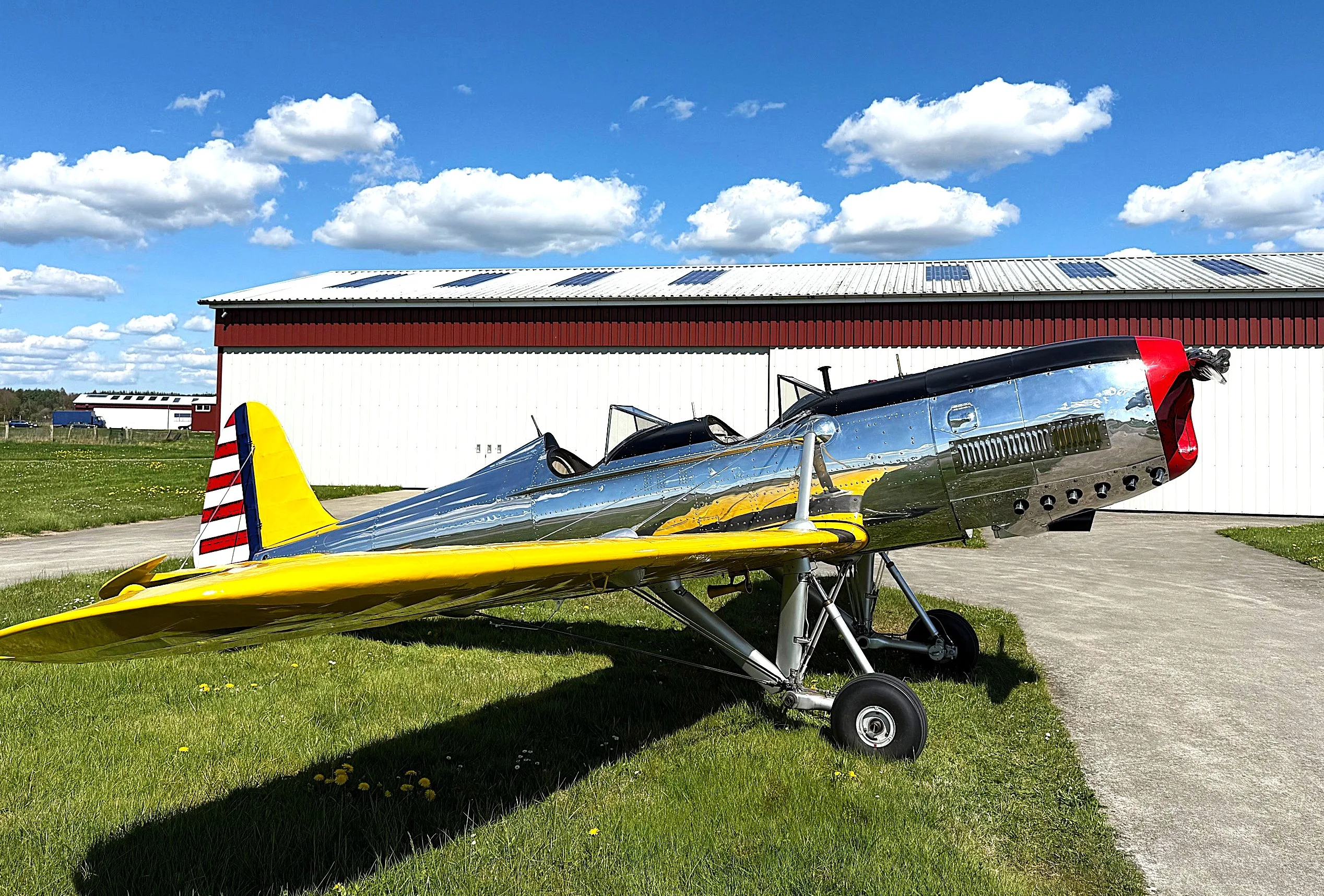 A shiny chrome and yellow vintage aircraft with a red nose and striped tail, parked on grass next to a white hangar under a blue sky with clouds.