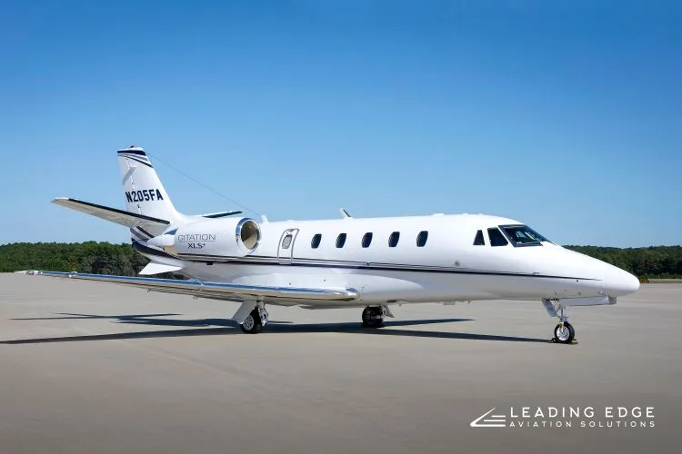 A white Cessna Citation XLS+ business jet with black and grey stripes parked on an tarmac under a clear blue sky.