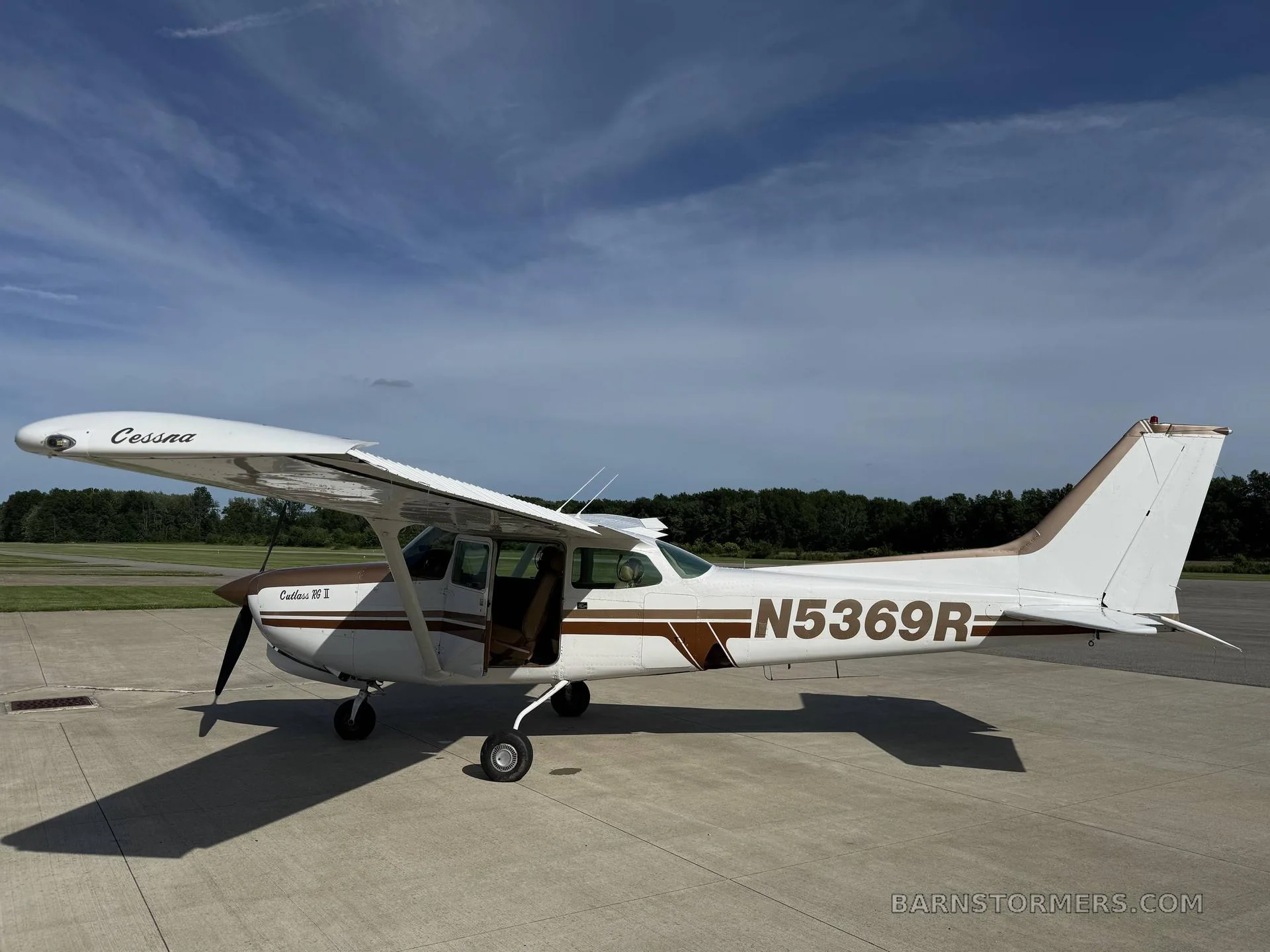 A white and brown Cessna Cutlass RG II (N5369R) is parked on an asphalt ramp with its door open, under a blue sky.