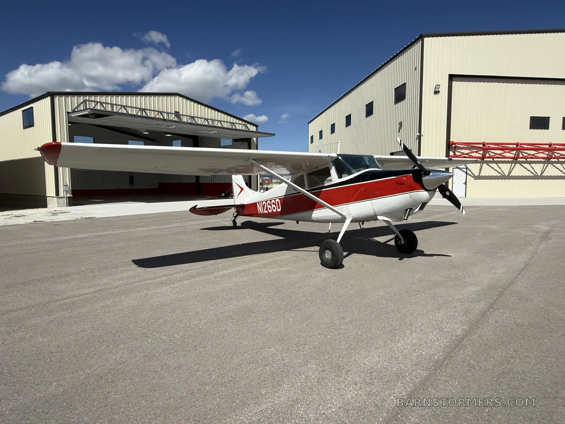 A red and white Cessna 170B (N1266D) is parked on an airfield, viewed from the front-right, with hangars in the background.